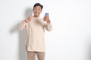 Portrait of young excited Asian muslim man in koko shirt holding mobile phone, taking picture of himself or selfie, saying hi and waving his hand. Social media concept. Isolated on white background photo