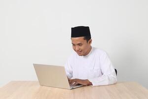retrato de emocionado asiático musulmán hombre en koko camisa con casquete trabajando en su ordenador portátil durante rápido en Ramadán mes. aislado imagen en blanco antecedentes foto