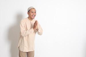 Portrait of attractive Asian muslim man in koko shirt with skullcap showing apologize and welcome hand gesture. Apology during eid mubarak. Isolated image on white background photo