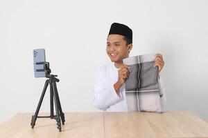 Portrait of excited Asian muslim man in koko shirt with skullcap promoting his product, sarung, on live streaming session. Online shopping with smartphone concept. Isolated image on white background photo