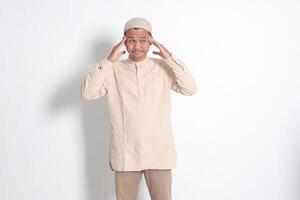 retrato de sufrimiento asiático musulmán hombre en blanco camisa teniendo un migraña, conmovedor su templo. dolor de cabeza enfermedad concepto. aislado imagen en blanco antecedentes foto