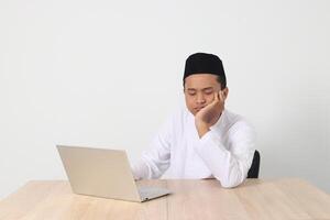 Portrait of tired Asian muslim man in koko shirt with skullcap working during fasting on ramadan month, feeling sleepy, yawning with hand covering mouth. Isolated image on white background photo