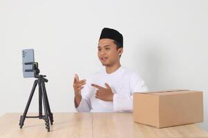 Portrait of excited Asian muslim man in koko shirt with skullcap promoting his product on live streaming session. Online shopping with smartphone concept. Isolated image on white background photo