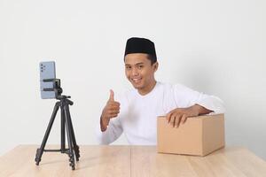 Portrait of excited Asian muslim man in koko shirt with skullcap promoting his product on live streaming session. Online shopping with smartphone concept. Isolated image on white background photo