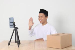 Portrait of excited Asian muslim man in koko shirt with skullcap promoting his product on live streaming session. Online shopping with smartphone concept. Isolated image on white background photo