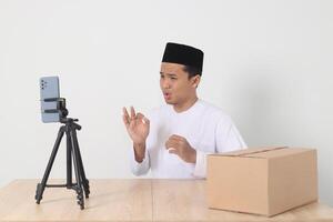 Portrait of excited Asian muslim man in koko shirt with skullcap promoting his product on live streaming session. Online shopping with smartphone concept. Isolated image on white background photo