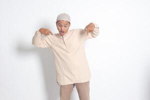 Portrait of shocked Asian muslim man in koko shirt with skullcap showing product and pointing with his hand and finger down. Advertising concept. Isolated image on white background photo