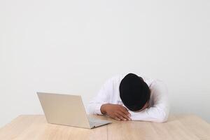 Portrait of excited Asian muslim man in koko shirt with skullcap working during fasting on ramadan month, feeling sleepy, sleeping at his table with laptop. Isolated image on white background photo