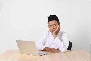 Portrait of tired Asian muslim man in koko shirt with skullcap working during fasting on ramadan month, feeling sleepy, yawning with hand covering mouth. Isolated image on white background photo