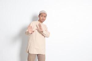 retrato de desagradable asiático musulmán hombre en koko camisa con casquete formando un mano gesto a evitar alguna cosa o malo cosas. aislado imagen en blanco antecedentes foto