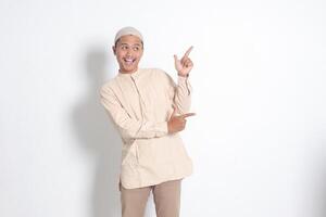 Portrait of shocked Asian muslim man in koko shirt with skullcap showing product and pointing with his hand and finger to the side. Advertising concept. Isolated image on white background photo