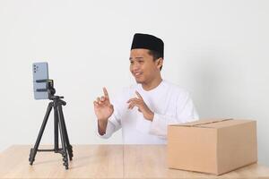 Portrait of excited Asian muslim man in koko shirt with skullcap promoting his product on live streaming session. Online shopping with smartphone concept. Isolated image on white background photo