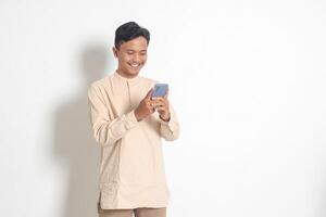 Portrait of young excited Asian muslim man in koko shirt holding mobile phone with smiling expression on face. Social media and internet concept. Isolated image on white background photo