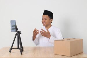 Portrait of excited Asian muslim man in koko shirt with skullcap promoting his product on live streaming session. Online shopping with smartphone concept. Isolated image on white background photo