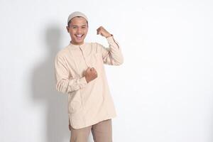 retrato de religioso asiático musulmán hombre en koko camisa con casquete levantamiento su puño, celebrando éxito. logro concepto. aislado imagen en blanco antecedentes foto