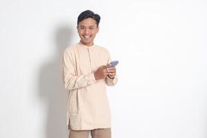 Portrait of young excited Asian muslim man in koko shirt holding mobile phone with smiling expression on face. Social media and internet concept. Isolated image on white background photo