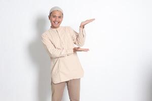 Portrait of shocked Asian muslim man in koko shirt with skullcap showing product and pointing with his hand and finger to the side. Advertising concept. Isolated image on white background photo