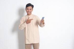 Portrait of young excited Asian muslim man in koko shirt holding mobile phone, pointing and showing product with finger. Social media concept. Isolated image on white background photo