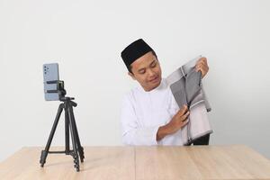Portrait of excited Asian muslim man in koko shirt with skullcap promoting his product, sarung, on live streaming session. Online shopping with smartphone concept. Isolated image on white background photo