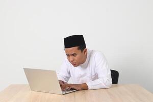 retrato de emocionado asiático musulmán hombre en koko camisa con casquete trabajando en su ordenador portátil durante rápido en Ramadán mes. aislado imagen en blanco antecedentes foto