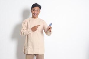 Portrait of young excited Asian muslim man in koko shirt holding mobile phone, pointing and showing product with finger. Social media concept. Isolated image on white background photo