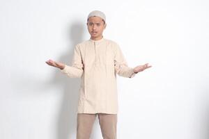 Portrait of confused Asian muslim man in koko shirt with skullcap spreading his hands sideways and holding two things, demonstrate products. Isolated image on white background photo