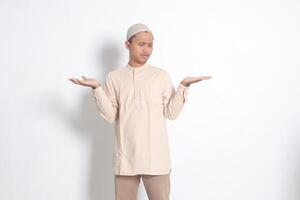 Portrait of confused Asian muslim man in koko shirt with skullcap spreading his hands sideways and holding two things, demonstrate products. Isolated image on white background photo