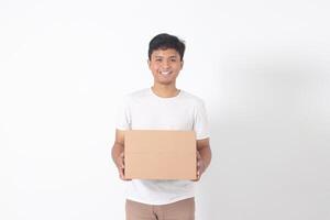 retrato de atractivo asiático hombre en blanco camiseta que lleva un cartulina caja. aislado imagen en blanco antecedentes foto