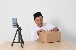 Portrait of excited Asian muslim man in koko shirt with skullcap promoting his product on live streaming session. Online shopping with smartphone concept. Isolated image on white background photo