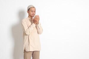 Portrait of religious Asian muslim man in koko shirt with skullcap praying earnestly with his hands raised. Devout faith concept. Isolated image on white background photo