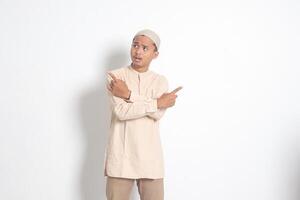 Portrait of excited Asian muslim man in koko shirt with skullcap pointing sideways, making choice, choosing between two objects, demonstrate product, crossed hands. Isolated image on white background photo