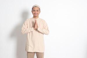 Portrait of attractive Asian muslim man in koko shirt with skullcap showing apologize and welcome hand gesture. Apology during eid mubarak. Isolated image on white background photo