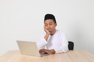 Portrait of tired Asian muslim man in koko shirt with skullcap working during fasting on ramadan month, feeling sleepy, yawning with hand covering mouth. Isolated image on white background photo