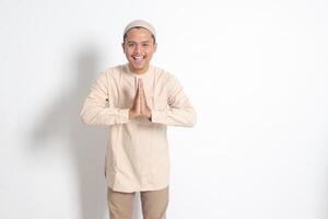 Portrait of attractive Asian muslim man in koko shirt with skullcap showing apologize and welcome hand gesture. Apology during eid mubarak. Isolated image on white background photo