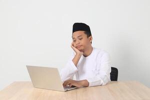 Portrait of tired Asian muslim man in koko shirt with skullcap working during fasting on ramadan month, feeling sleepy, yawning with hand covering mouth. Isolated image on white background photo