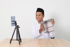 Portrait of excited Asian muslim man in koko shirt with skullcap promoting his product, sarung, on live streaming session. Online shopping with smartphone concept. Isolated image on white background photo