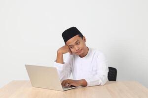 Portrait of tired Asian muslim man in koko shirt with skullcap working during fasting on ramadan month, feeling sleepy, yawning with hand covering mouth. Isolated image on white background photo