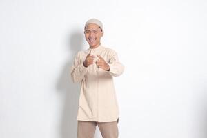 Portrait of shocked Asian muslim man in koko shirt with skullcap showing product and pointing with his hand and finger forward to the camera. Advertising concept. Isolated image on white background photo