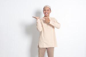 Portrait of shocked Asian muslim man in koko shirt with skullcap showing product and pointing with his hand and finger to the side. Advertising concept. Isolated image on white background photo