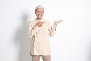 Portrait of shocked Asian muslim man in koko shirt with skullcap showing product and pointing with his hand and finger to the side. Advertising concept. Isolated image on white background photo