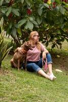 Smiling woman with sunglasses hugging her dog, seated in a park area. photo