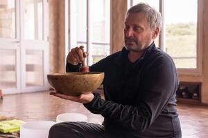 A mature man playing a bronze bowl, seated and illuminated by light streaming through the window. photo