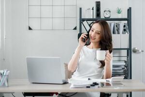 Relaxed businesswoman enjoys a coffee while having a phone conversation at her office desk with a laptop. photo