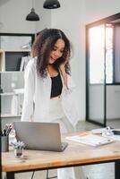 Businesswoman in a relaxed posture chats on the phone, her expression one of pleasure and engagement, in her modern office with a laptop open before her. photo