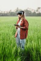 Woman holds a tablet and wears a virtual reality headset, standing in a lush green field with distant trees. photo