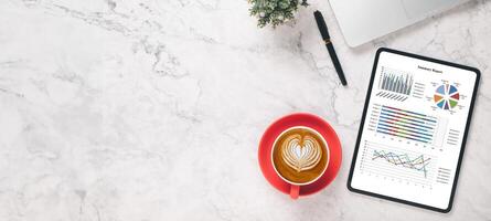 Angulo alto ver de un profesional espacio de trabajo presentando un en forma de corazon latté Arte café y un tableta mostrando vistoso negocio analítica cartas foto