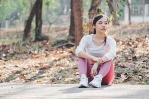 Woman runner resting after workout session on sunny morning. Female fitness model sitting on street along pond in city. Female jogger taking a break from running workout. photo