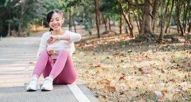 Joyful young woman sitting on a park path, looking at her smartwatch, enjoying a break after a workout session surrounded by nature. photo