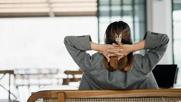 Rear view of a businesswoman taking a break at her workspace, with hands clasped behind her head and laptop on the table. photo