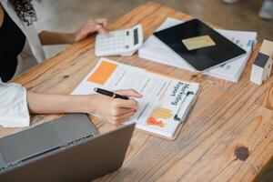 enfocado mujer de negocios utilizando un calculadora y revisando financiero informes, rodeado por varios trabajo documentos en su escritorio. foto